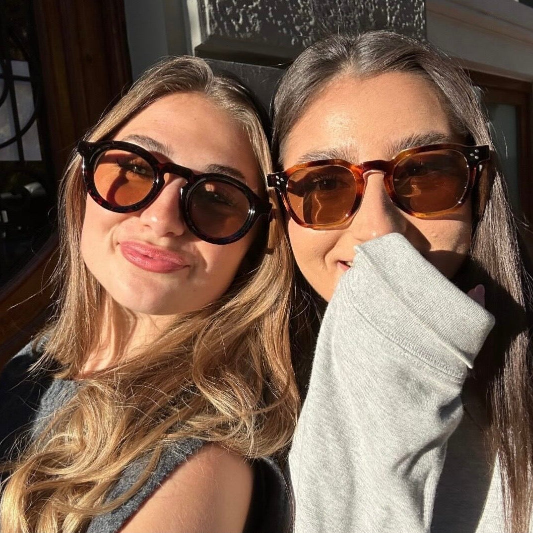 Two women wearing sunglasses with a top-down view of one of them blowing her nose.